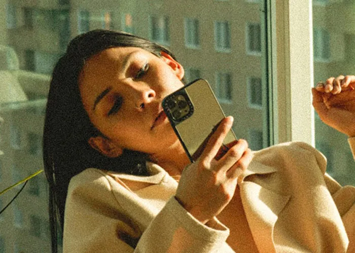 Young woman using a smartphone near a window, reflecting on how she exposed her cheating partner.