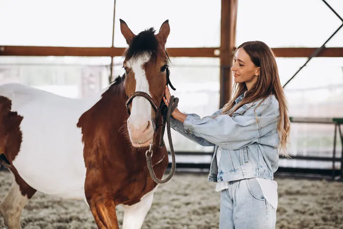 Young woman in denim jacket gently holding a brown and white horse in an indoor stable, demonstrating romanticized experience