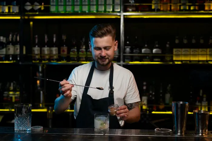 Bartender in a black apron preparing a cocktail behind the bar, representing things folks only romanticize.
