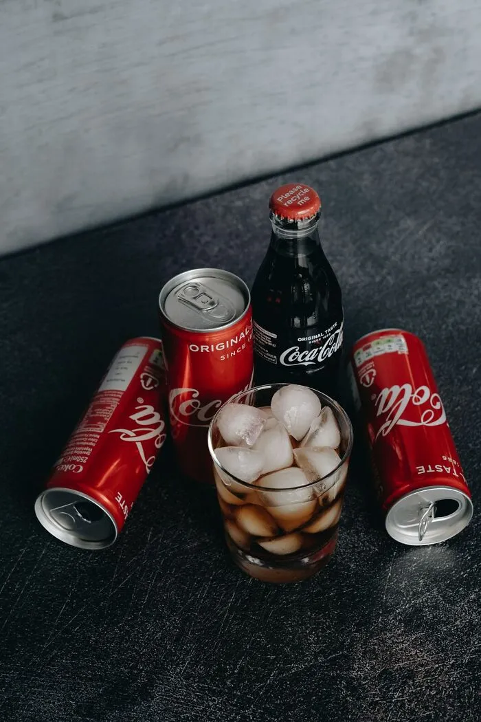Cans and bottle of Coca-Cola with a glass of ice on a dark surface representing restaurant food beverages.