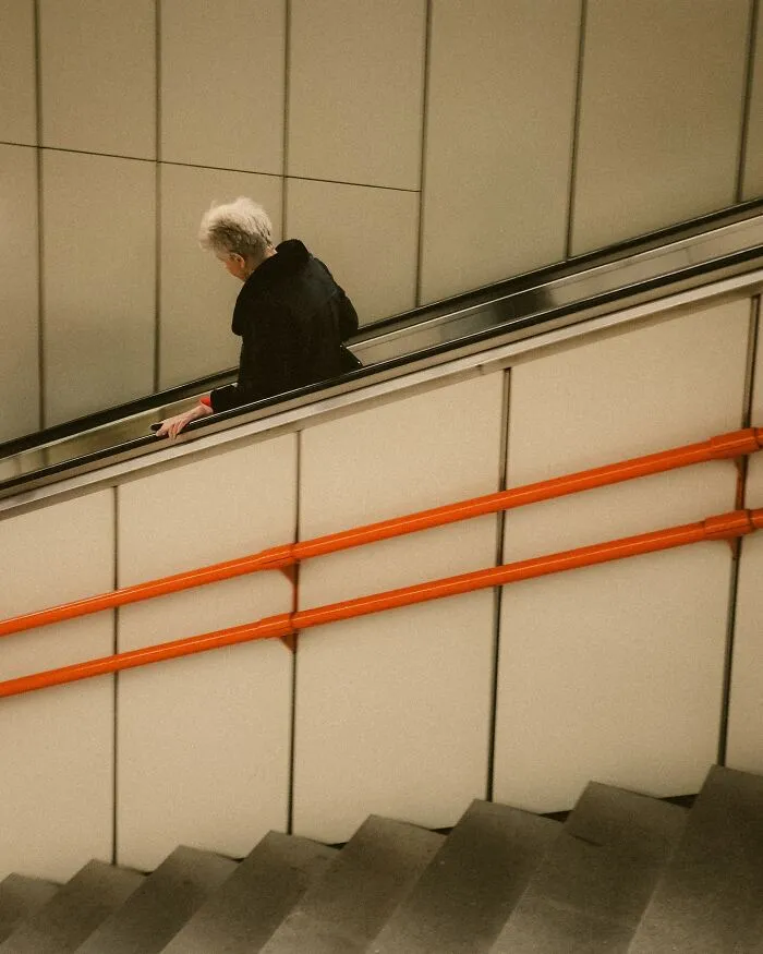 Elderly person in black coat on escalator with orange handrails in urban street photography style by Helsinki photographer