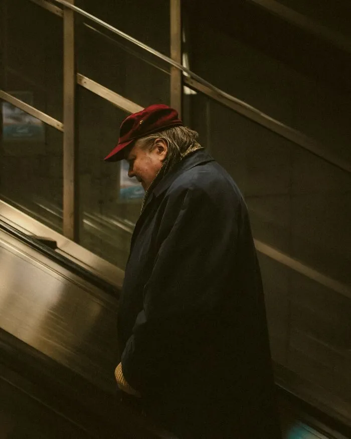 Elderly person in a red hat and dark coat standing on an escalator in an urban setting, captured in a street photo.