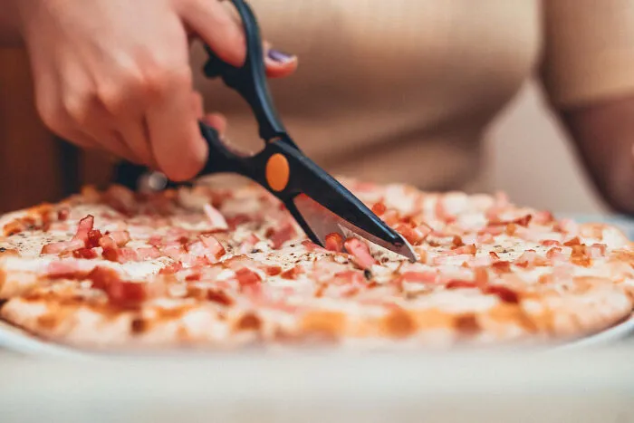 Hand using scissors to cut a pizza, illustrating behaviors Americans reveal non-Americans do that are like breaking spaghetti.