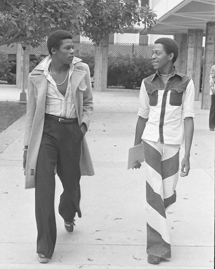 Two stylish young men walking outdoors, wearing vintage 1970s fashion in a fascinating historical photo.