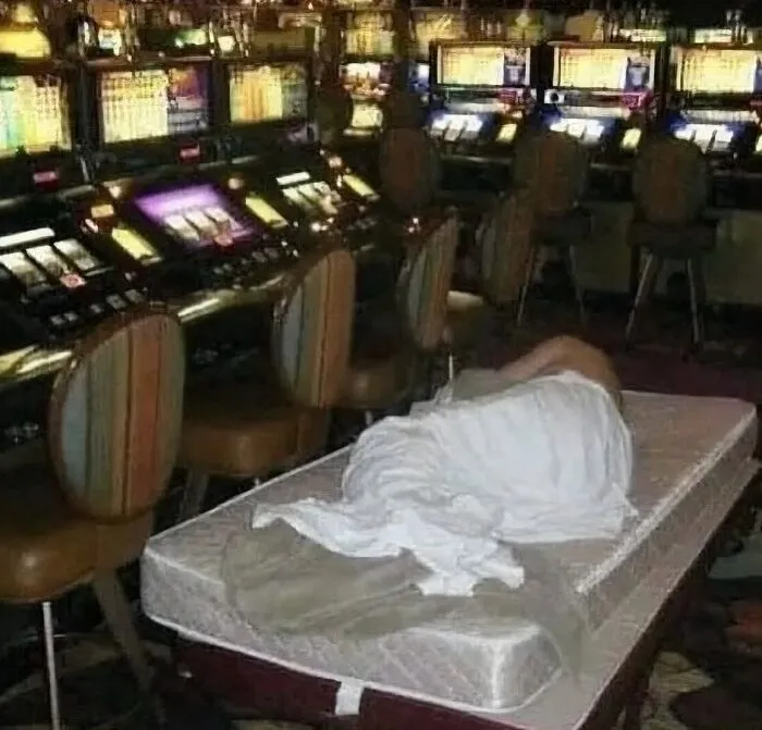Person covered with a blanket lying on a mattress in a dimly lit casino with slot machines and empty chairs around.