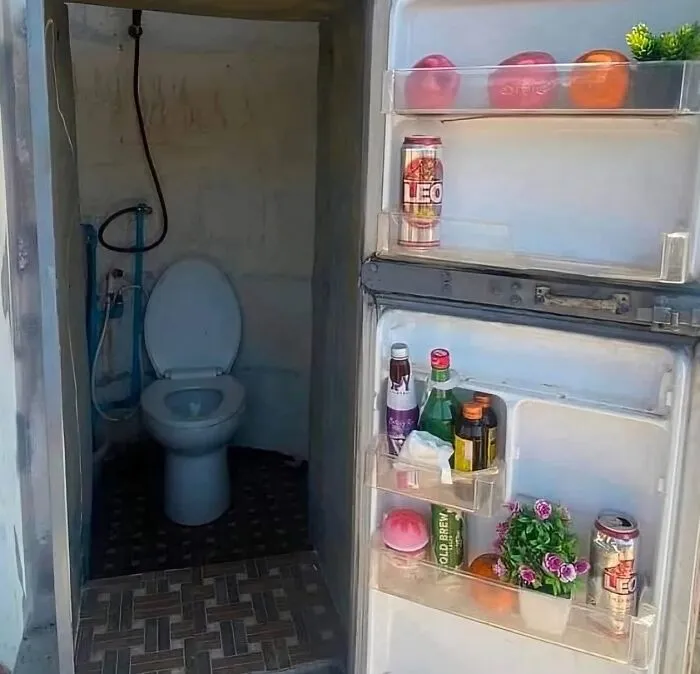 Open fridge door revealing beverages and flowers inside, with a toilet visible directly behind it, creating an uncomfortable scene.