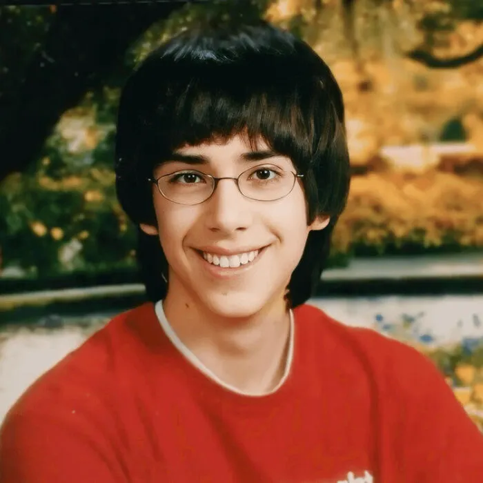Smiling young person with glasses wearing red shirt, posing outdoors with blurred fall foliage background in disturbing photos series.