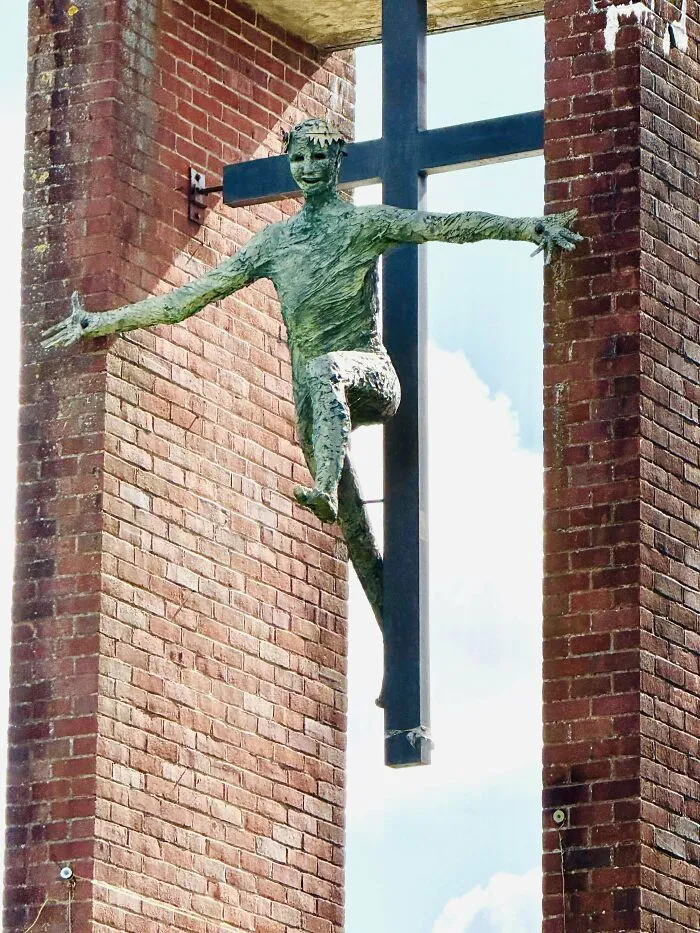 Disturbing photo of a creepy green statue with outstretched arms hanging between brick walls and a large black cross in the background.