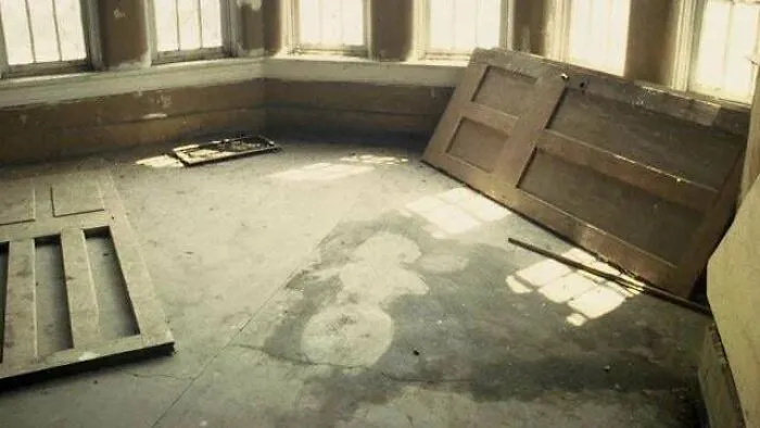 Abandoned room with broken doors and disturbing stain resembling a figure on the floor in a dimly lit space.