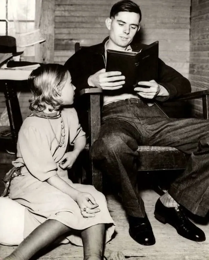 Black and white photo of a man reading a book to a young girl in a rustic room, evoking disturbing vintage vibes.