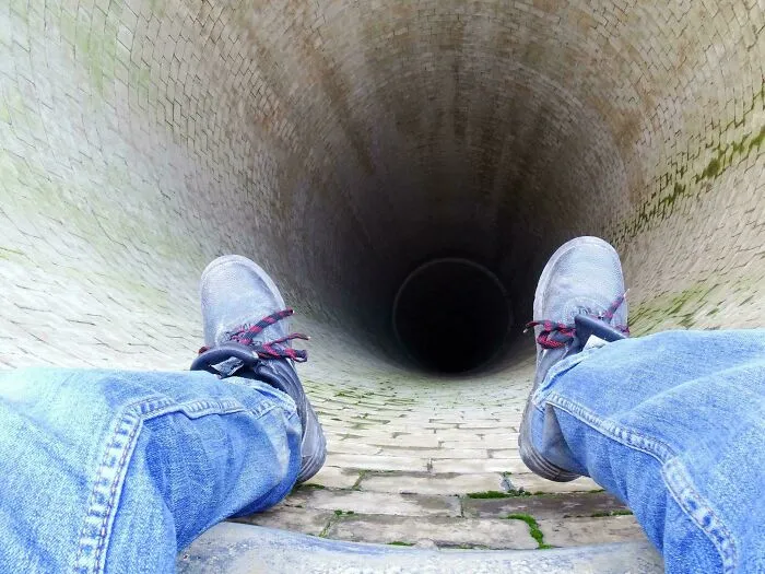 Piernas con pantalones de mezclilla y zapatos encima de un pozo profundo y oscuro, imagen aterradora e inquietante.