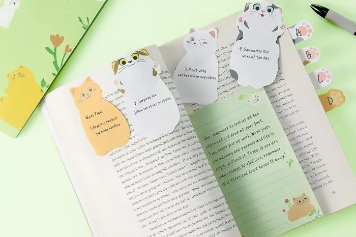 Cat-shaped office supplies including sticky notes and page markers arranged on an open book and green background.