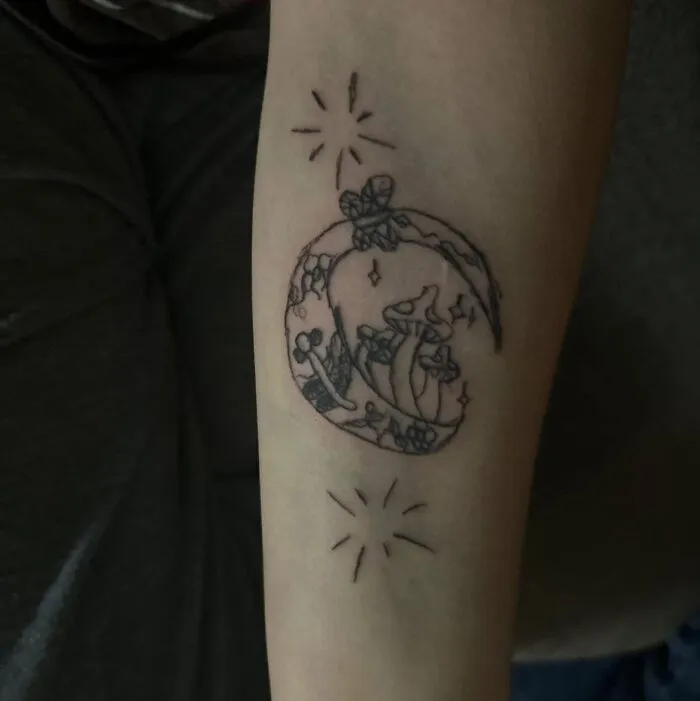 Black ink tattoo on forearm featuring a crescent moon with mushrooms and stars, an example of hilariously bad tattoos.
