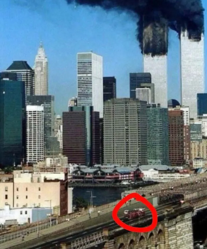 Historic photo of New York City skyline with Twin Towers burning and emergency vehicles highlighted on bridge in foreground.