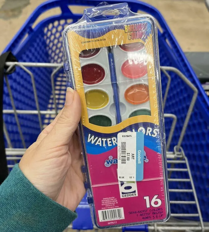 Hand holding a new 16-color watercolor paint set with price tag in a thrift store shopping cart.
