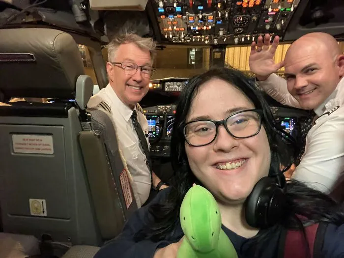 Smiling passenger with headphones holding a toy in a cockpit with pilots, capturing a wholesome moment overcoming fear of flying.