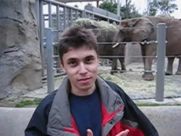 Young man at the zoo making humorous gestures, elephants visible in the background, capturing internet lies and absurd humor.
