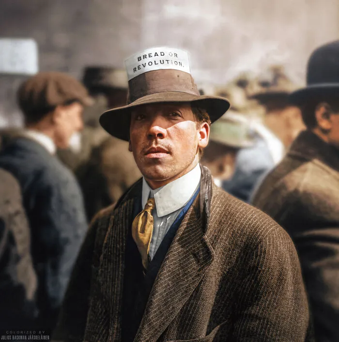 Colorized history pic of a man wearing a bread or revolution sign on his hat at a crowded gathering.