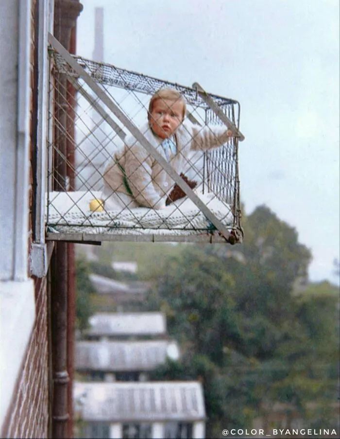 Child in a metal cage balcony outside a building, a rare interesting colorized history pic showing past child safety measures.