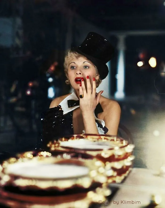 Woman in a black top hat and gloves with bright red nails, captured in an interesting colorized history pic setting.