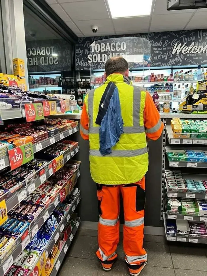 Hombre con chaleco y ropa de trabajo con pieza de ropa colgada en la espalda en tienda, foto divertida con humor cotidiano.