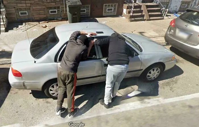 Two people caught in a shenanigan leaning into a car on a street, captured on Google Maps.