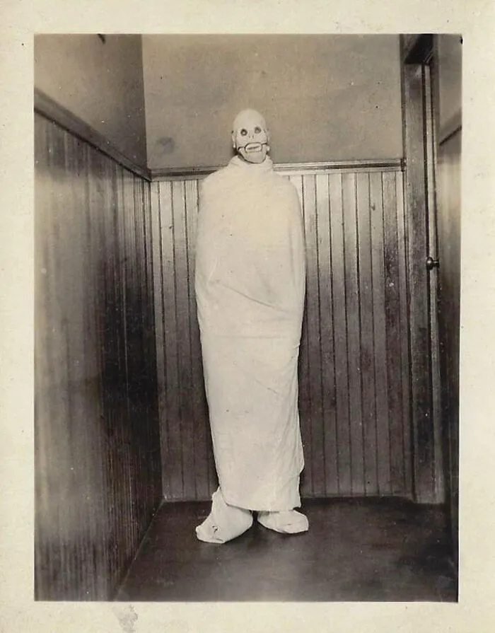 Foto antigua en blanco y negro de persona con disfraz inquietante envuelta en sábana en esquina de habitación pequeña.