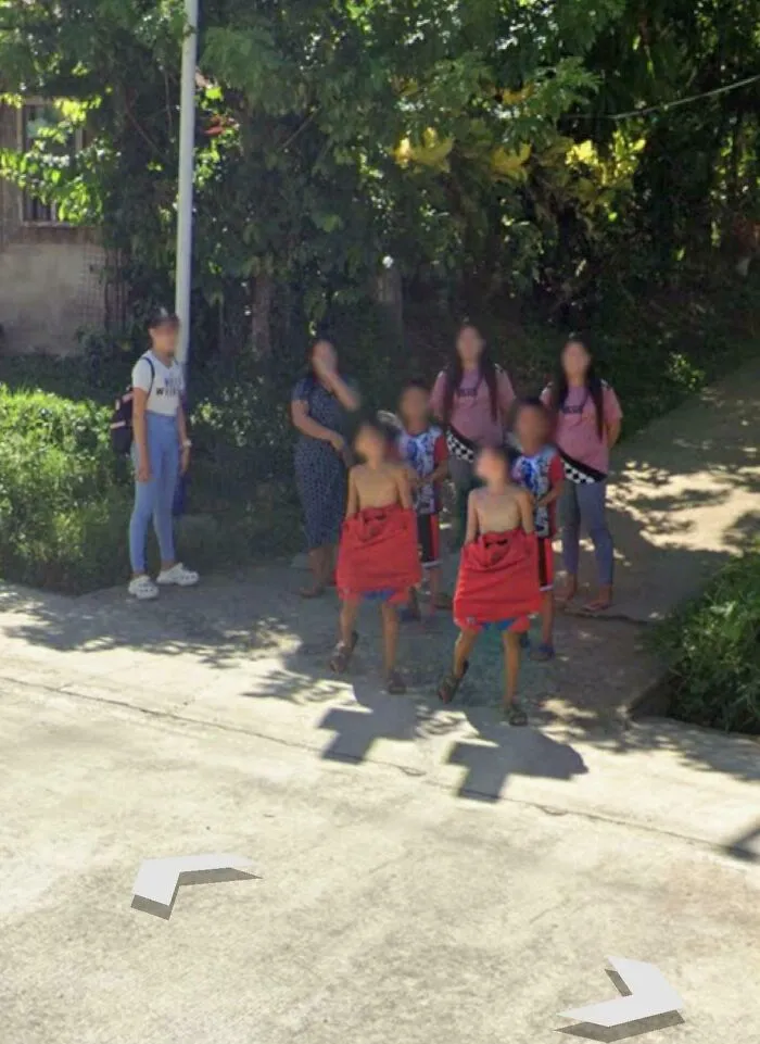 Group of people including children and adults caught in unusual moment on a street, captured in Google Maps shenanigans.