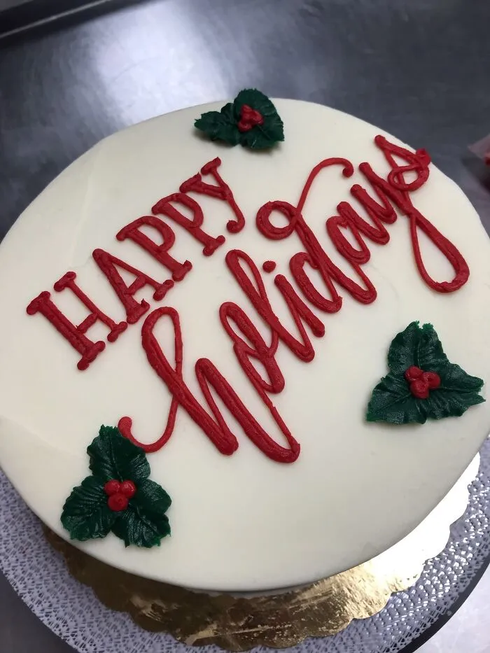 Cake with perfect handwriting example in red icing saying Happy Holidays with holly decorations on white frosting.