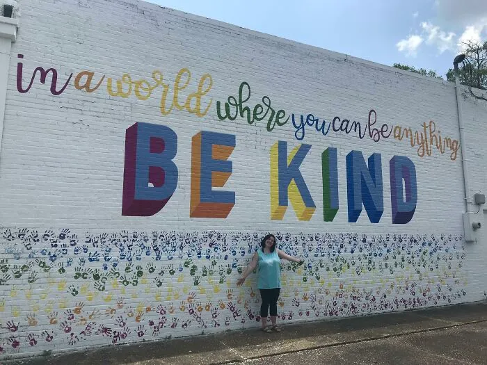 Colorful mural with perfect handwriting example saying be kind on white brick wall with person standing below it outdoors
