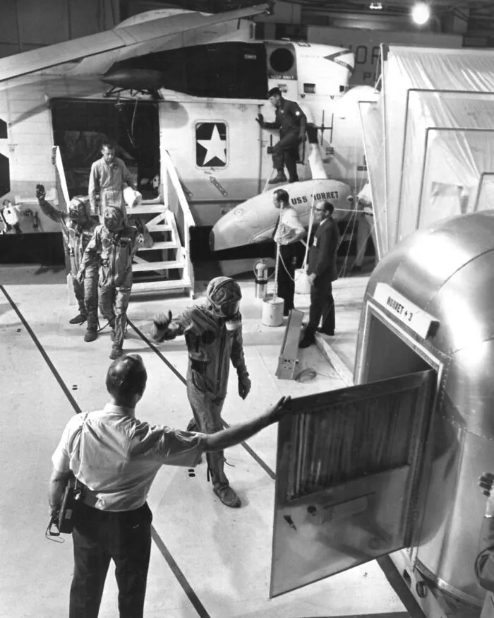 Apollo mission astronauts in training suits preparing for a simulation inside a spacecraft module with support crew nearby.