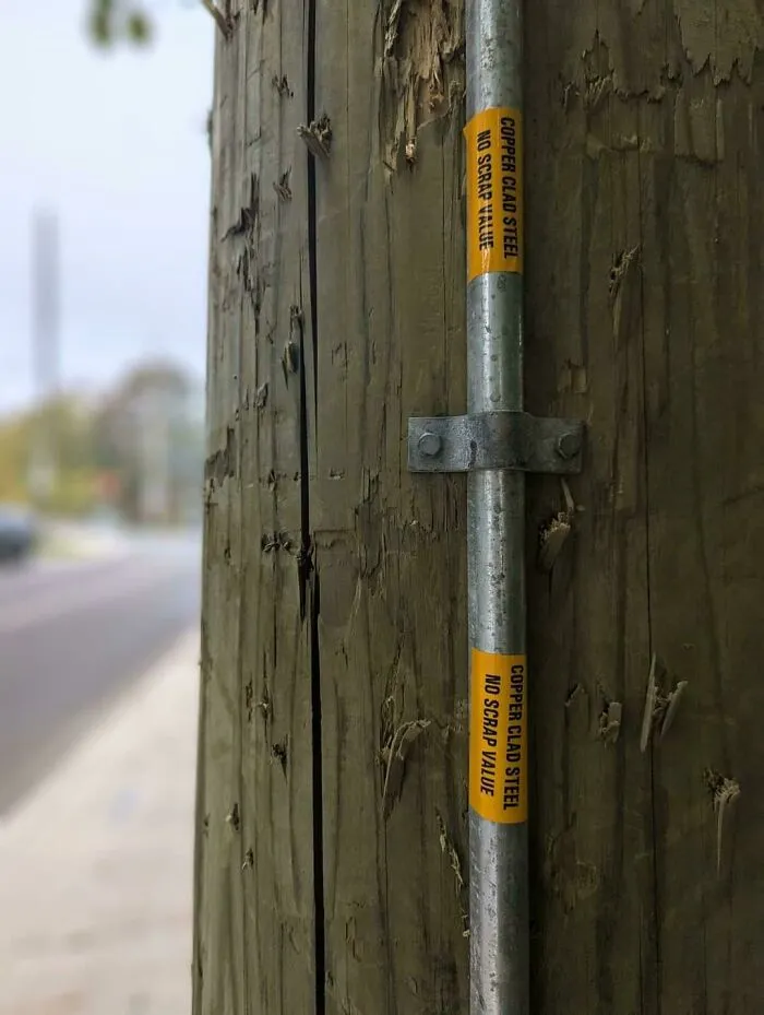 Close-up of a wooden utility pole with metal conduit labeled to prevent copper clad steel scrap theft near a blurred street.