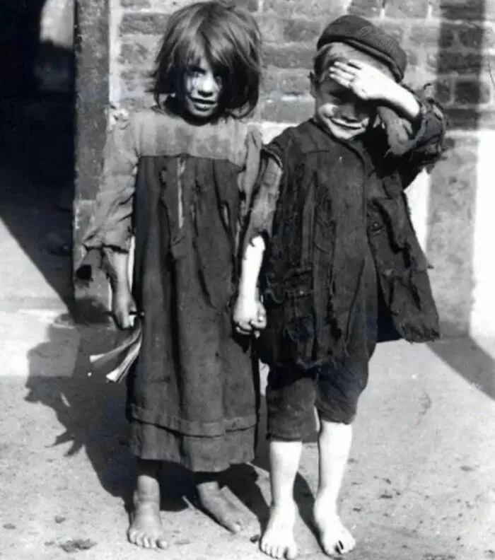 Two barefoot children in worn clothing holding hands in a black and white historical photo depicting past hardships.