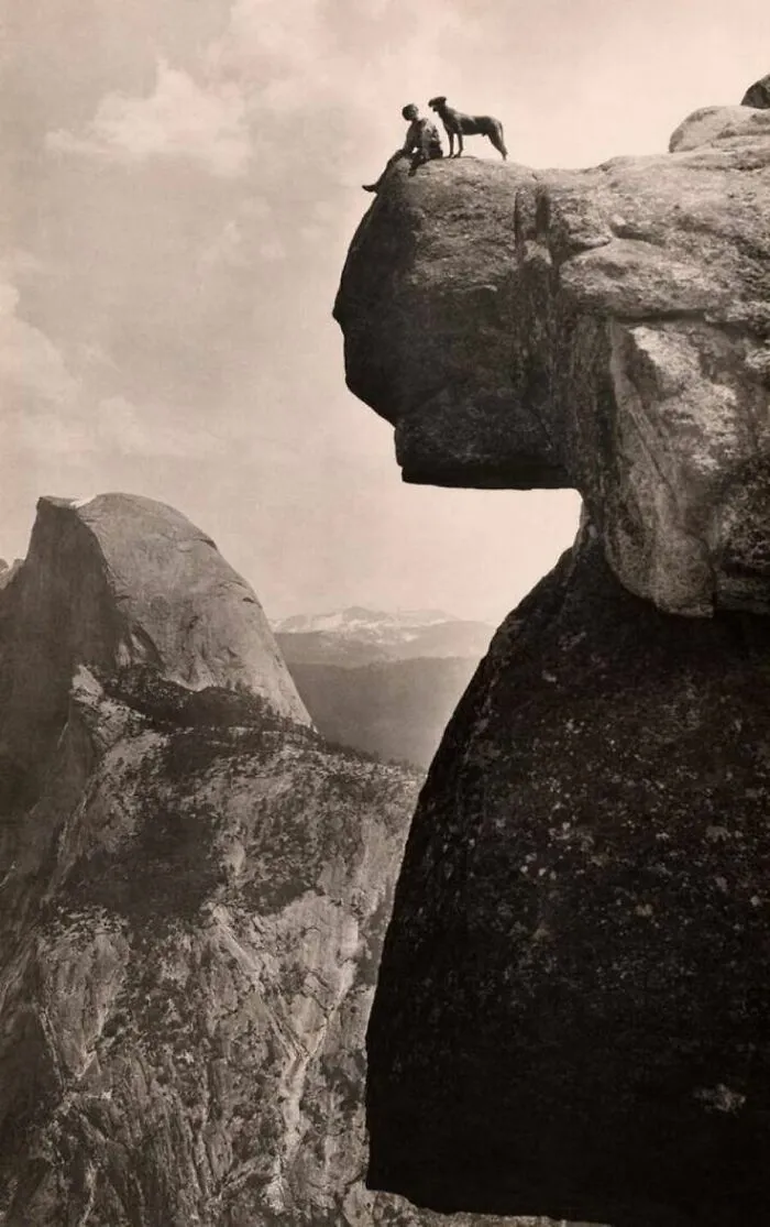 Vintage historical photo of a person and dog sitting on a cliff edge with vast mountain landscape below.