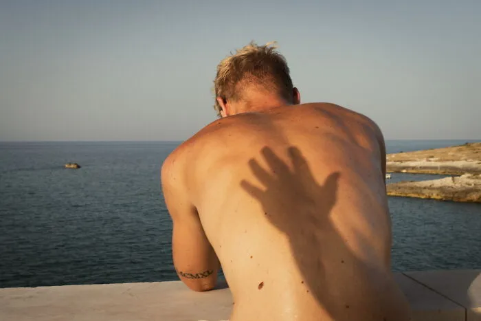 Shirtless man leaning on ledge by the sea with a shadow of a hand perfectly timed on his back in street photos style.