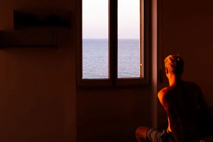 Man sitting indoors looking out a window at the sea during sunset, captured in a perfectly-timed street photo.