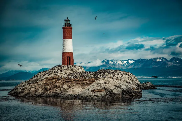 Lighthouse on rocky island surrounded by sea birds and mountains, representing scary places around the world for brave travelers