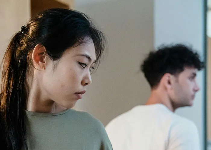 Young woman looking upset in the foreground while man faces away in a room, illustrating called off engagements theme.
