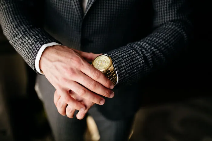 Man in a patterned suit flaunting wealth by prominently displaying a luxury gold watch on his wrist.