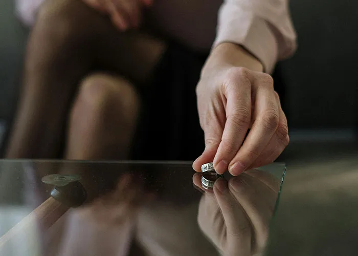 Person sitting on a couch, holding an engagement ring near a glass table, symbolizing called off engagements.