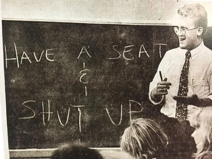 Profesor en aula con pizarra mostrando curiosa foto histórica que refleja métodos de enseñanza poco convencionales.