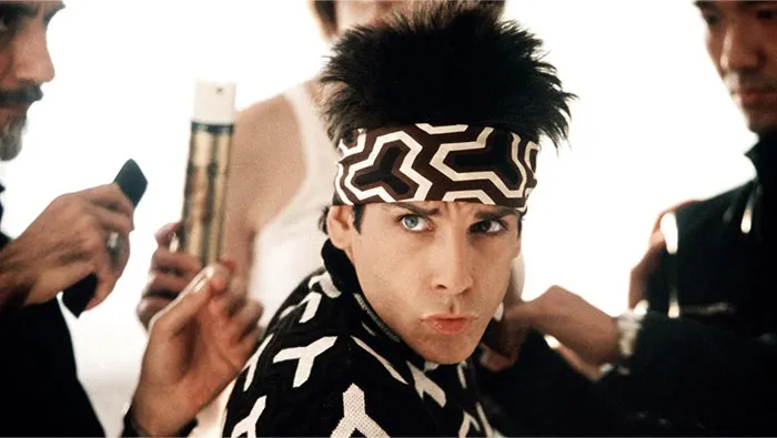 Actor wearing a patterned headband and jacket, surrounded by stylists preparing him for a photo shoot on set.