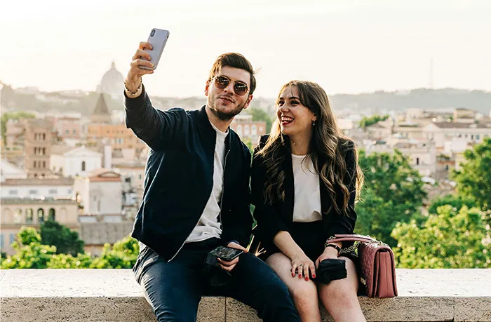 A young couple sitting outdoors in a cityscape, taking a selfie together and smiling on a sunny day.