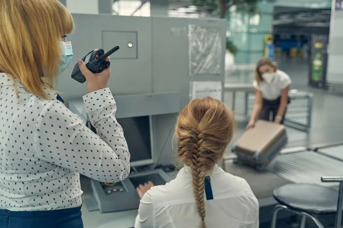 Airport security staff wearing masks inspect luggage and communicate using a walkie-talkie during baggage screening.