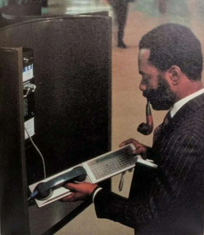 A Man Checks His E-Mail Over A Public Pay Telephone Using A Panasonic Rl-P4001 Acoustic Coupler Dial-Up Modem Attached To A Panasonic Rl-H1400 Hhc (Hand-Held Computer) In The Early 1980s