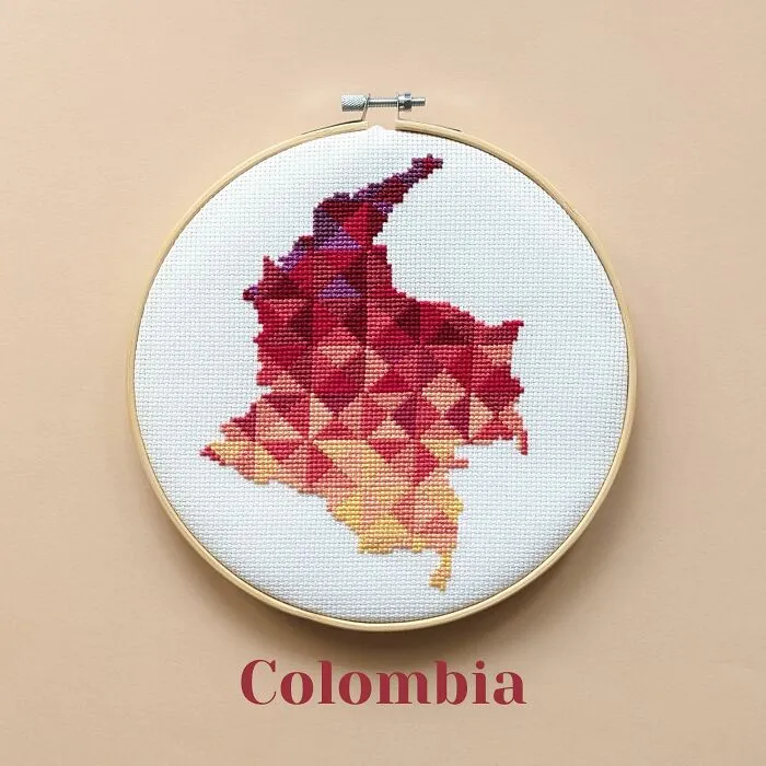 Cross-stitched map of Colombia in warm red and orange shades displayed in a wooden embroidery hoop on beige background.