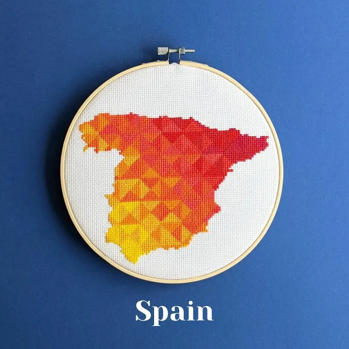 Cross-stitched map of Spain in warm gradient colors displayed in an embroidery hoop on a blue background.