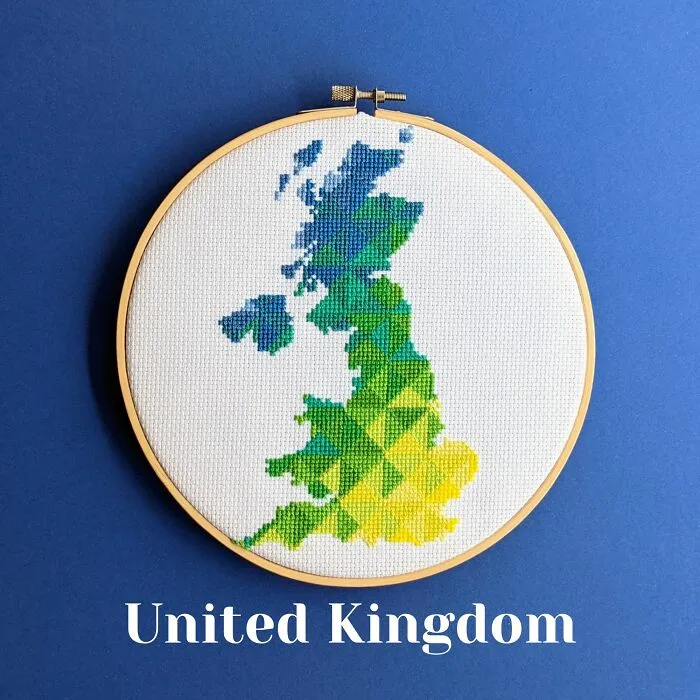 Cross-stitched map of the United Kingdom in green, blue, and yellow thread inside an embroidery hoop on a blue background.