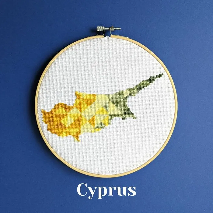 Cross-stitched map of Cyprus with geometric patterns in yellow and green shades in an embroidery hoop on blue background