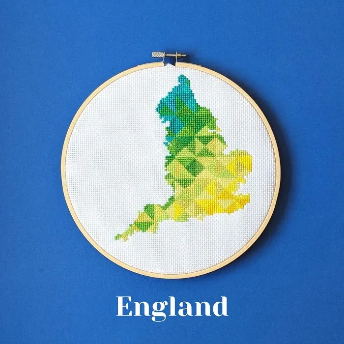 Cross-stitched map of England in green, blue, and yellow threads displayed in an embroidery hoop on a blue background.